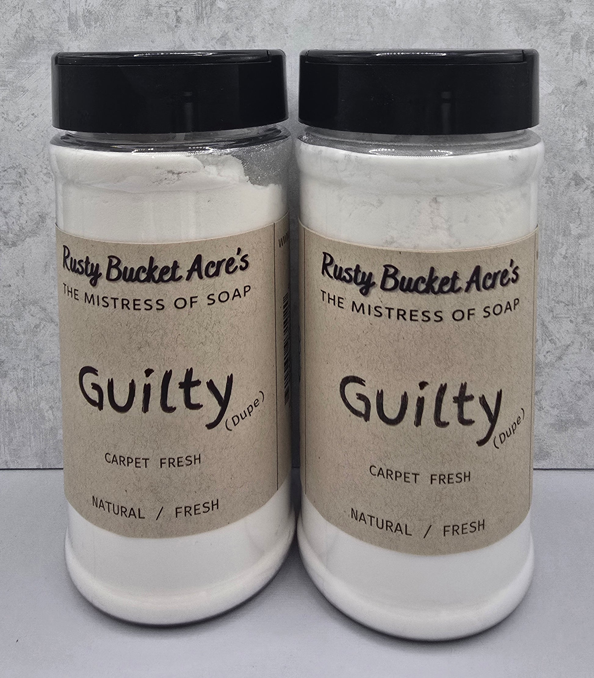 Two clear plastic jars of Rusty Bucket Acre's 'Guilty (Duple)' Carpet Fresh natural soap powder, side-by-side against a gray textured background.
