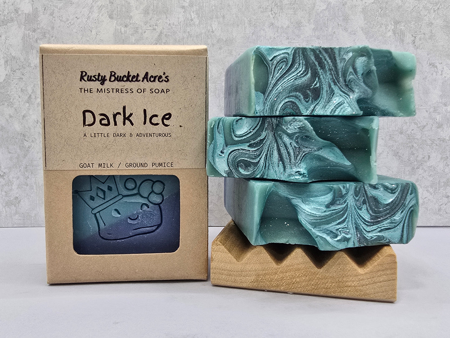 Three marbled soap bars in a teal and gray swirl pattern are stacked on a small wooden stand next to their kraft paper packaging box labeled 'Rusty Bucket Acre's Dark Ice'.