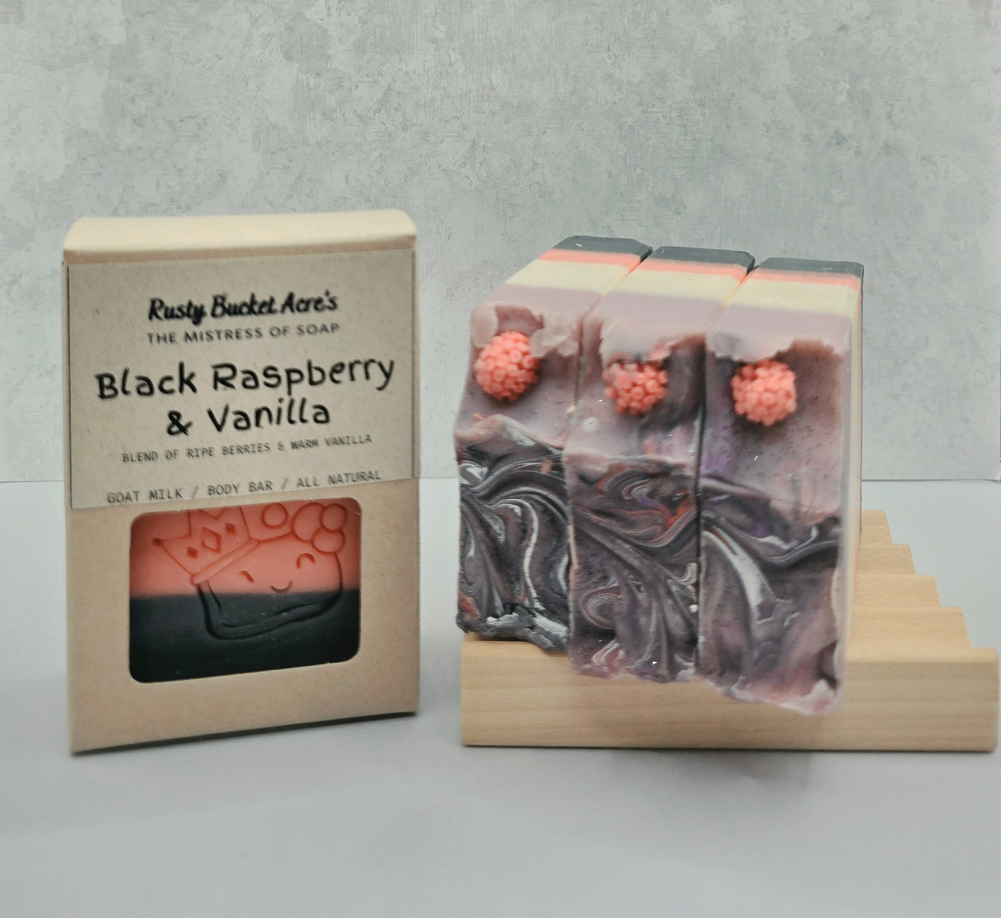A boxed bar of soap and three unwrapped soap bars displayed on a small wooden stand. The box reads 'Rusty Bucket Acre's Black Raspberry & Vanilla,' and the soaps show a dark marbled body with pink raspberry accents and small raspberry decorations on top, set against a light gray textured background.