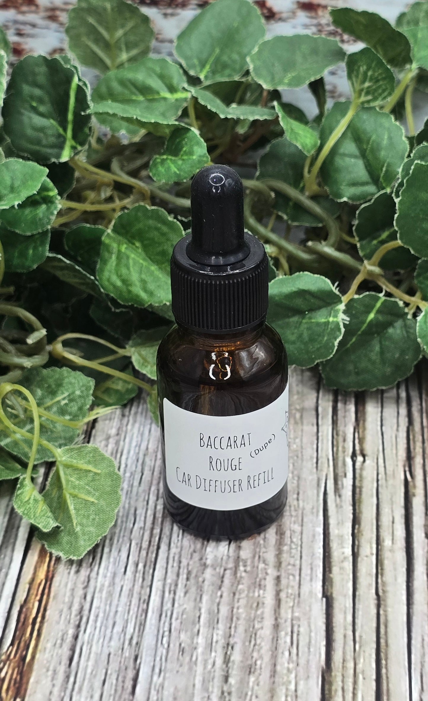 Bottle of Baccarat Rouge diffuser oil with a dropper on a wooden surface with green leaves.