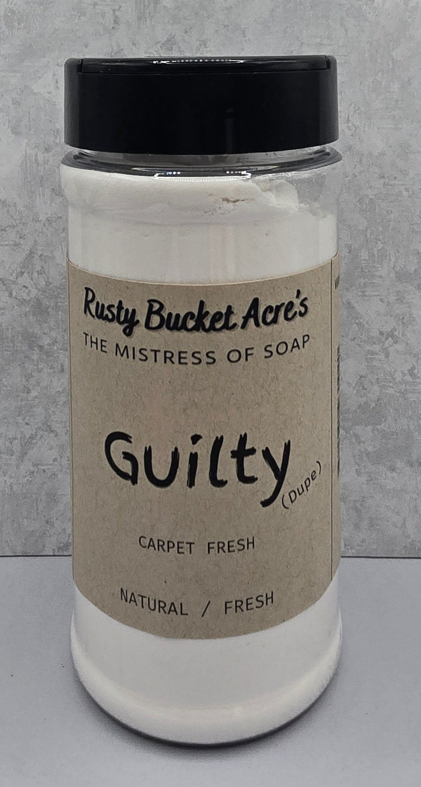 A white plastic jar of Rusty Bucket Acre's 'Guilty (Dupe)' carpet fresh soap with a black lid and a beige kraft paper label, set against a gray textured background.