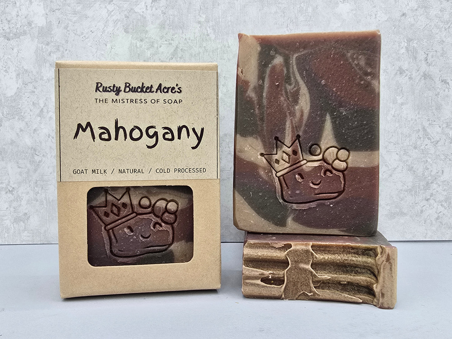 A handmade bar of soap named 'Mahogany' by Rusty Bucket Acre's, shown next to its kraft paper packaging box and a textured gold-colored soap tool resting on top of the bar.