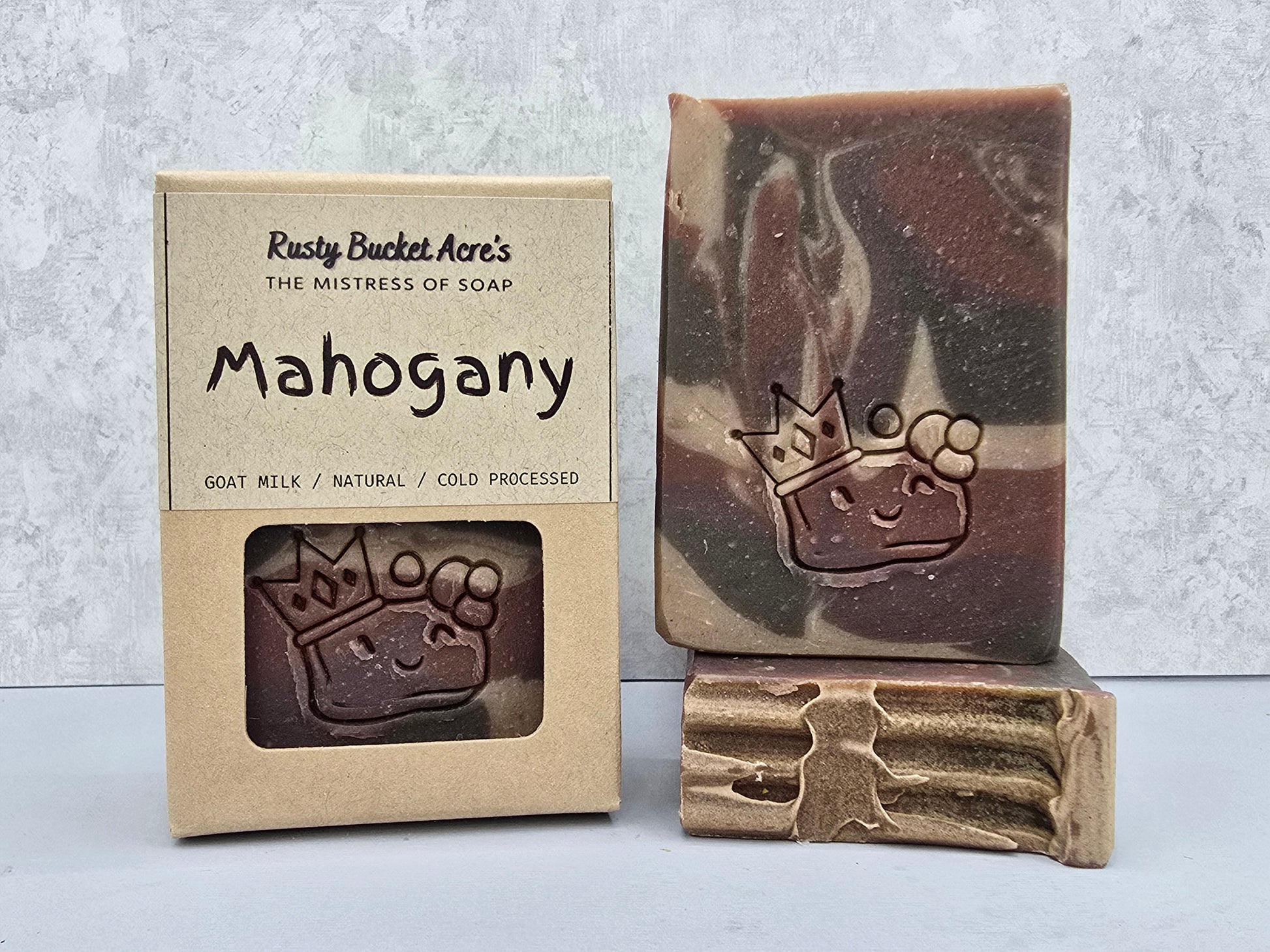 A handmade bar of soap named 'Mahogany' by Rusty Bucket Acre's, shown next to its kraft paper packaging box and a textured gold-colored soap tool resting on top of the bar.