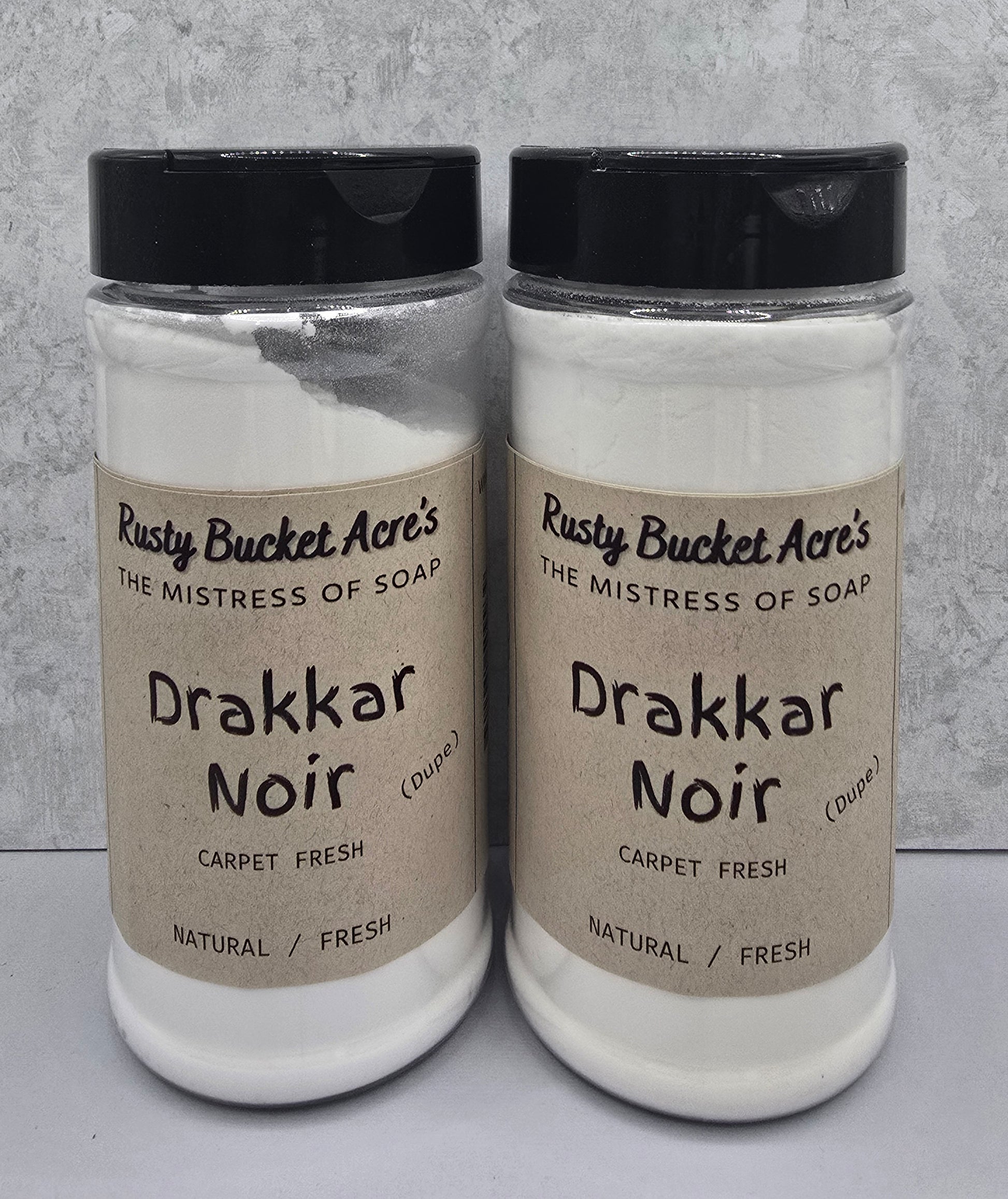 Two clear plastic jars of Rusty Bucket Acre's Drakkar Noir carpet fresh soap with black lids and beige labels, set against a gray textured background.