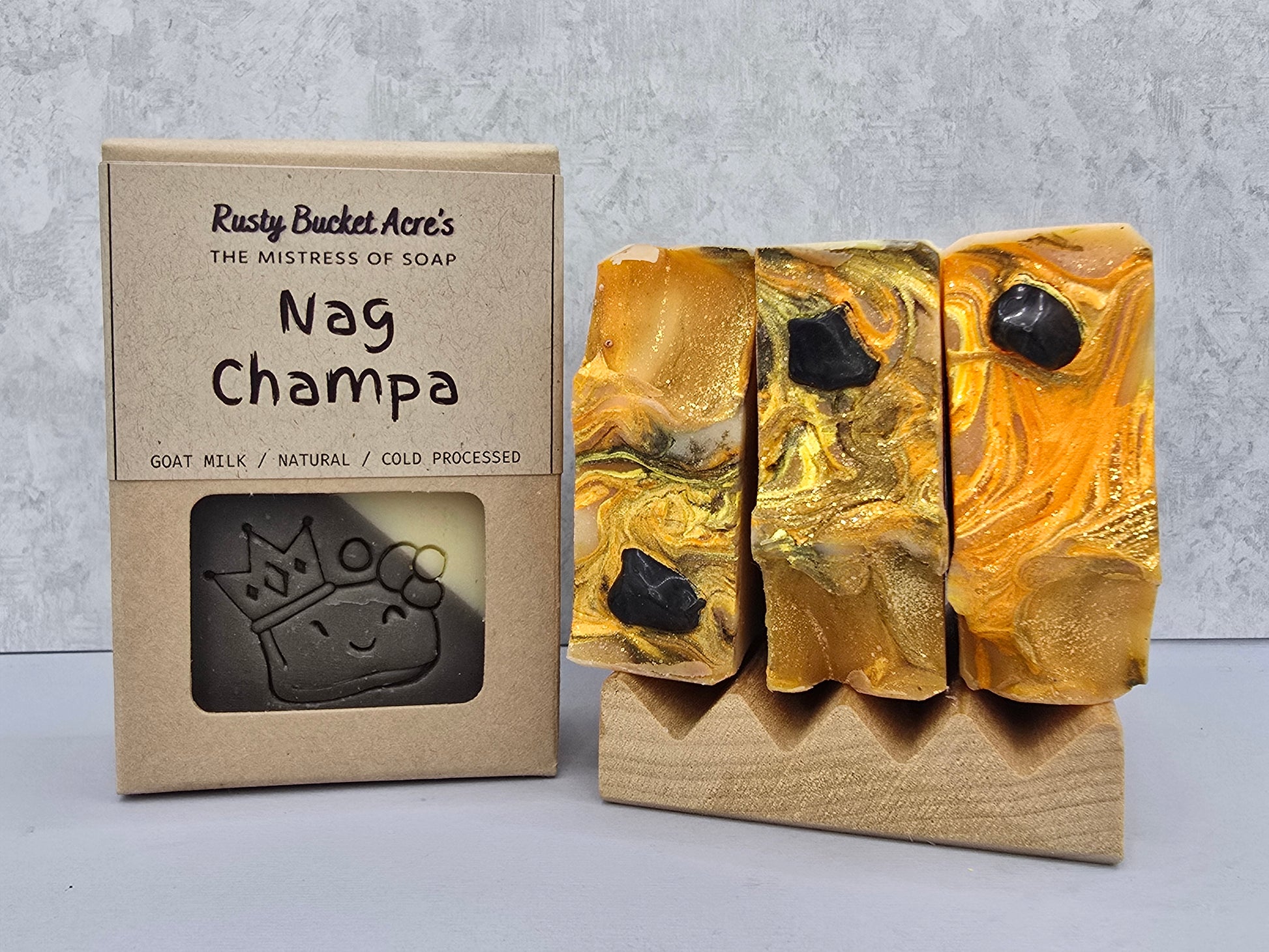 Three gold marbled soap bars with black accents on a wooden stand next to their kraft paper packaging box labeled 'Nag Champa' by Rusty Bucket Acre's.