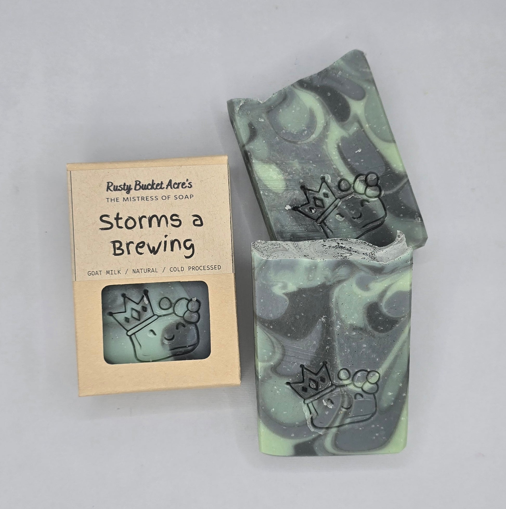 A bar of marbled soap with black, gray, and mint green swirls, next to its kraft paper packaging box labeled 'Storms a Brewing' by Rusty Bucket Acre's.