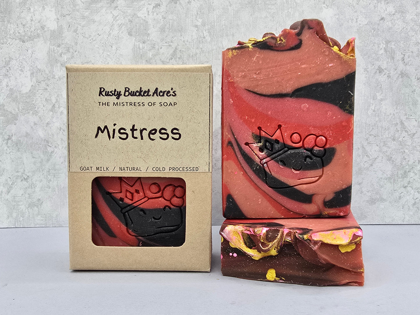 A handmade marbled soap bar labeled 'Mogg' stands next to its kraft paper packaging box, which reads 'Rusty Bucket Acre's The Mistress of Soap Mistress'. The soap features swirls of coral pink, dark brown, and black with small gold flecks, set against a light gray textured background.